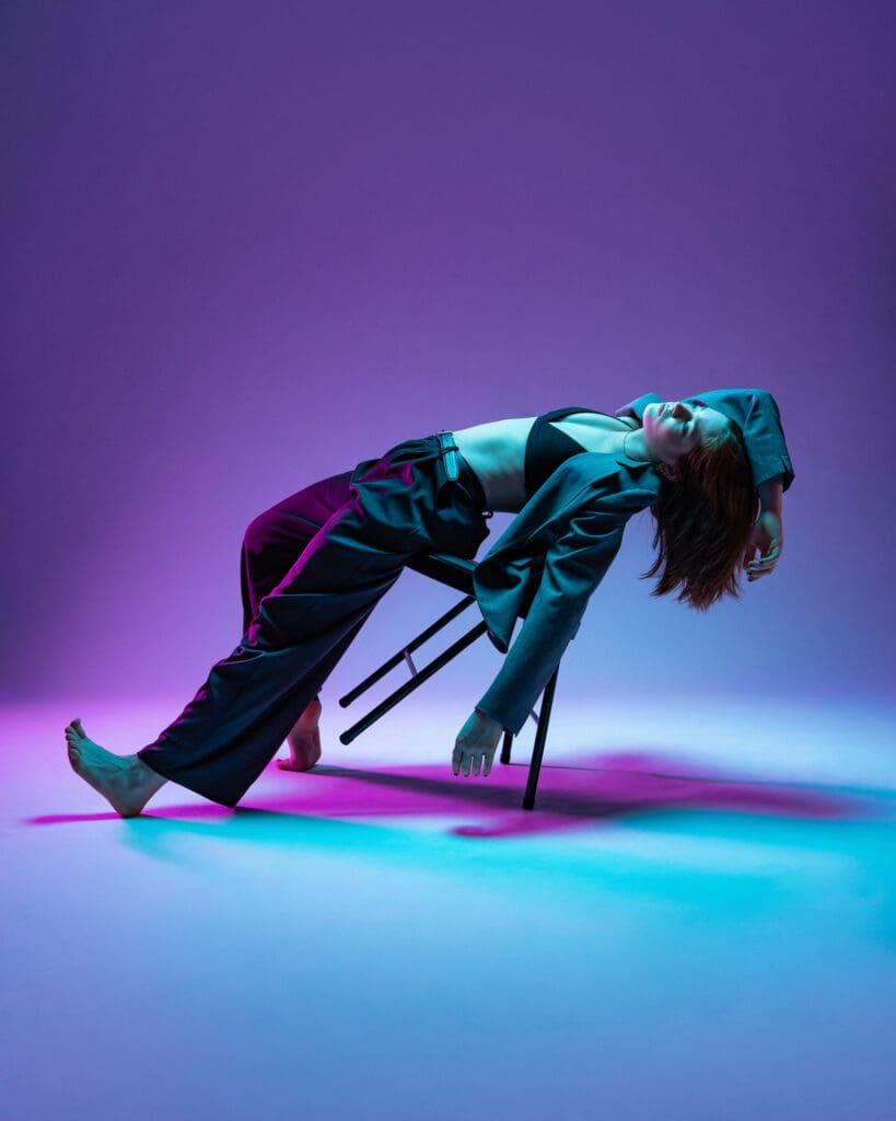 Contemporary dancer leaning back in chair with purple lighting overhead.