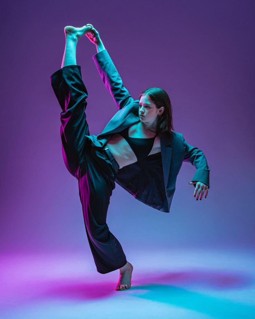 Contemporary dancer lifting leg in extension under purple and cyan lighting.