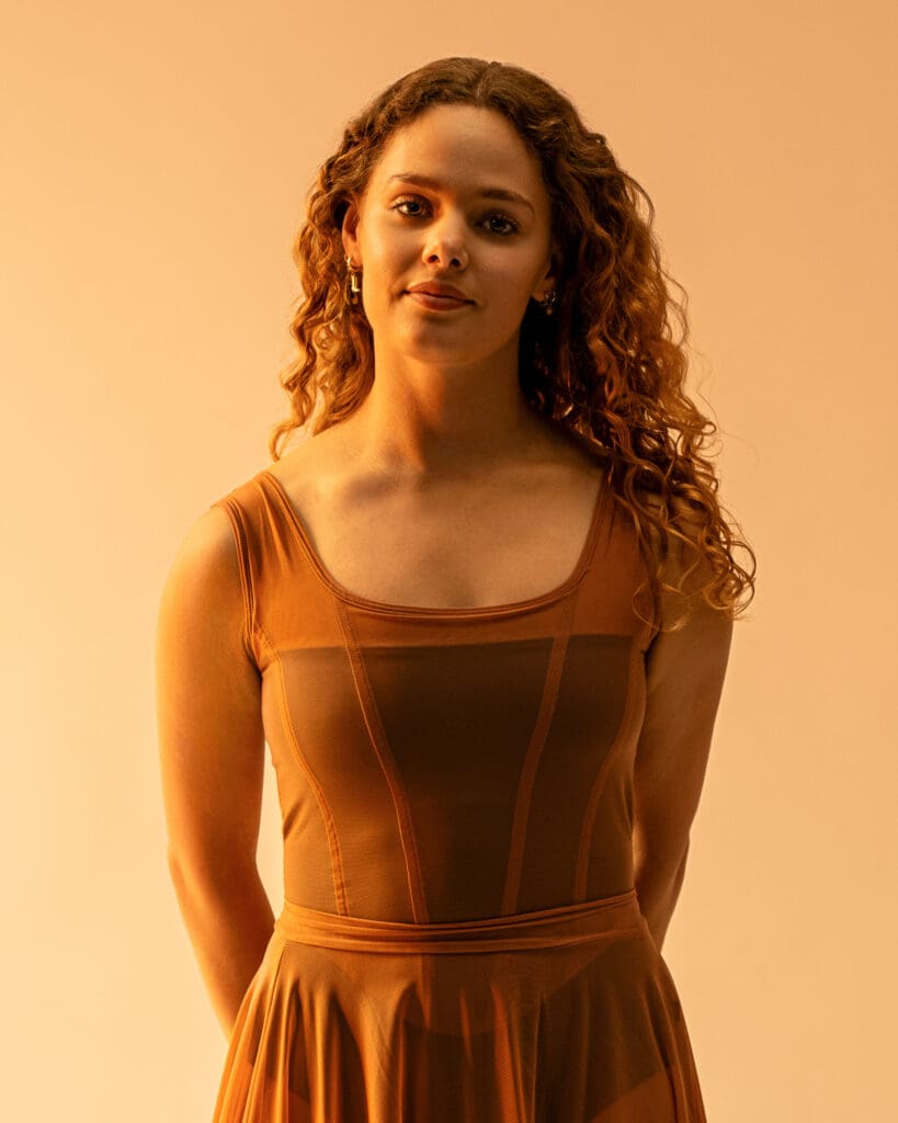 Young woman in orange dress in gold lighting gently smiles and looks at camera.