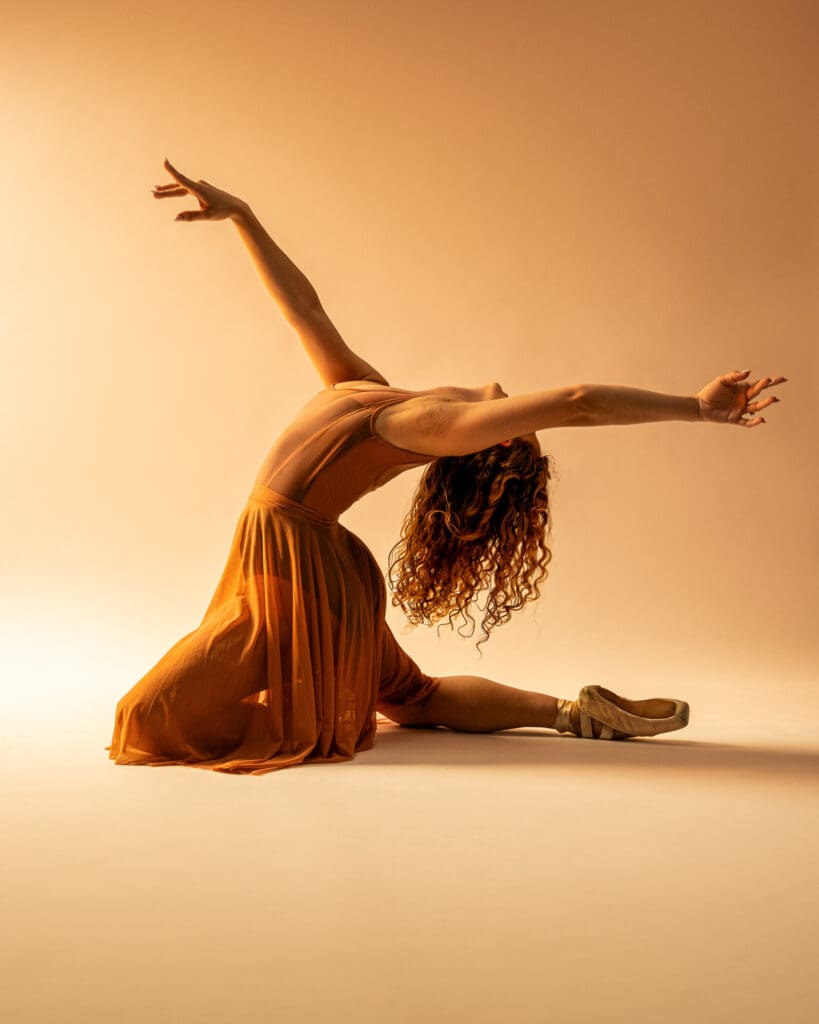 Ballet dancer performs contemporary improvisation on floor in gold lighting and orange dress.