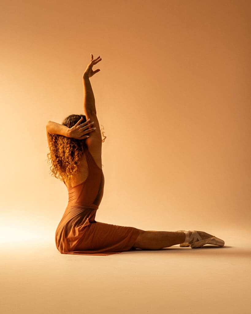 Ballet dancer on floor archers her back up and reaches to ceiling in gold lighting. 