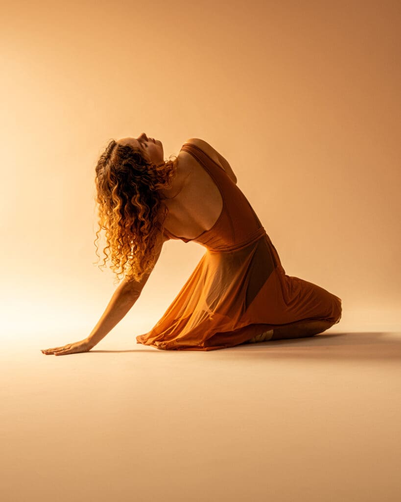 Dancer in golden light sits on floor on knees and leans back.
