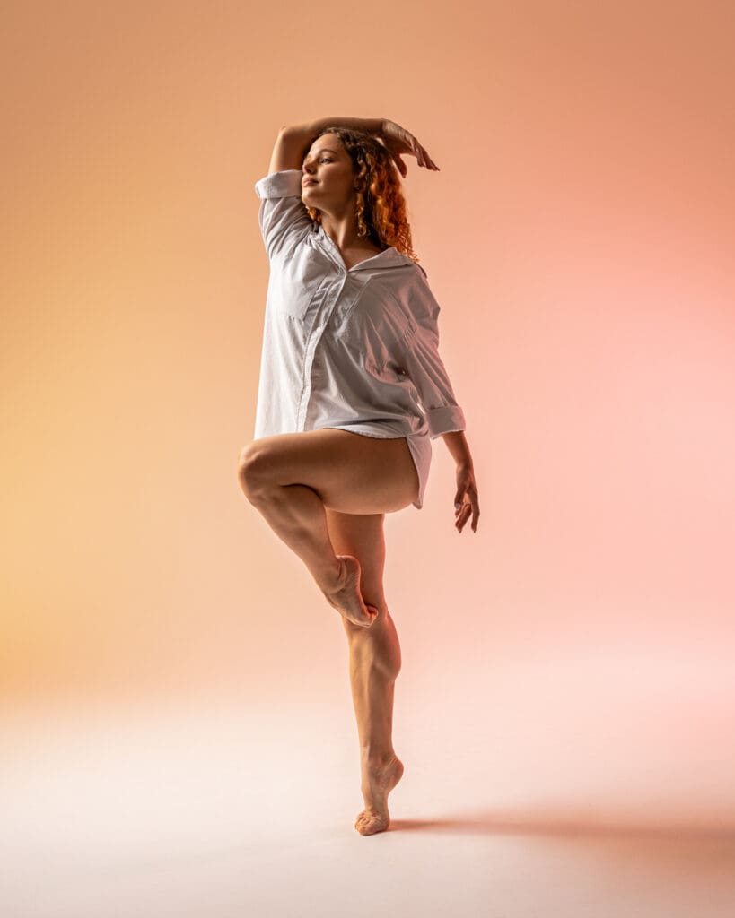 Dancer in white shirt and peach lighting performs passe on releve with arm draped over head.