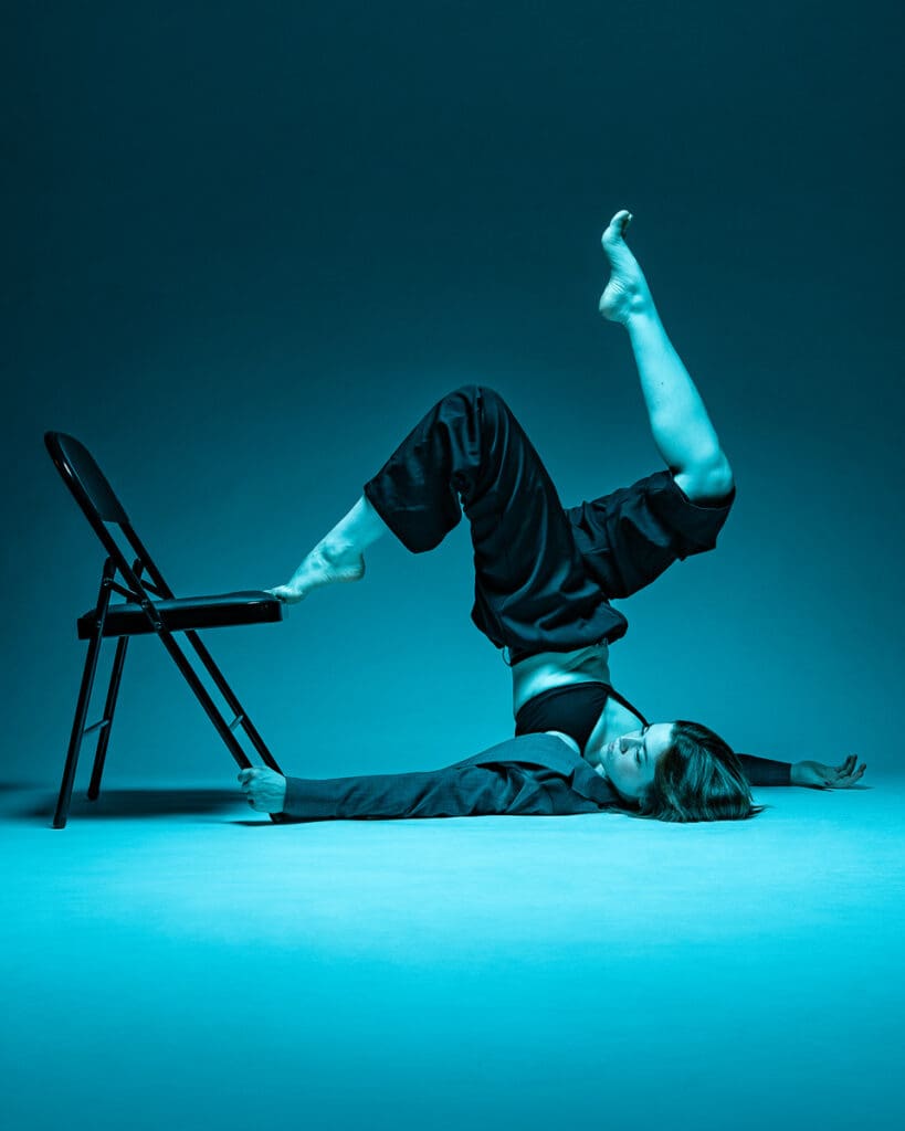 Dancer lying on the floor with one leg extended into a chair prop, teal lighting during a San Antonio contemporary dance photoshoot