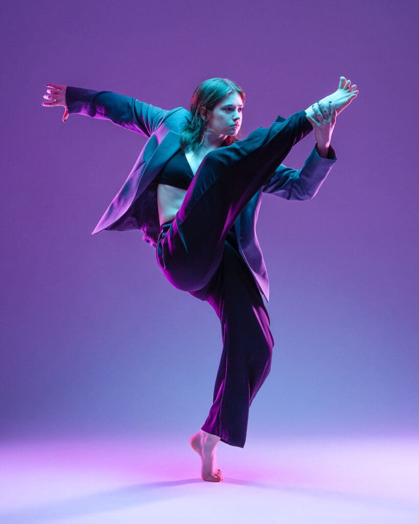 Contemporary dancer in a black blazer holding a high leg extension under purple studio lighting, photographed by a San Antonio dance photographer.