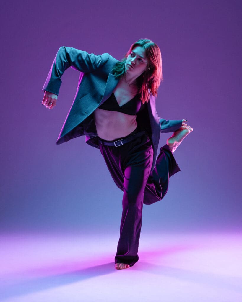 Contemporary dancer balanced on one leg in an open blazer with the other leg bent behind her, purple and teal lighting, photographed by a San Antonio dance photographer.
