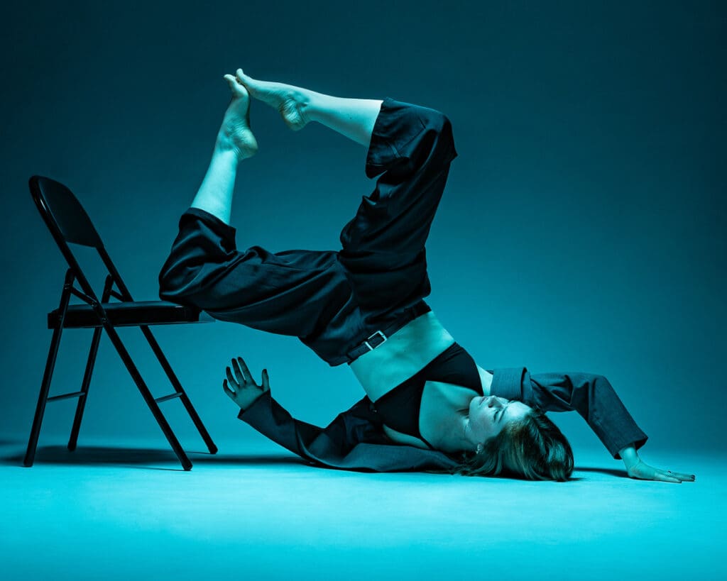A dancer exploring a contemporary floor pose with a chair under teal studio lighting.  Photographed by a San Antonio dance photographer.