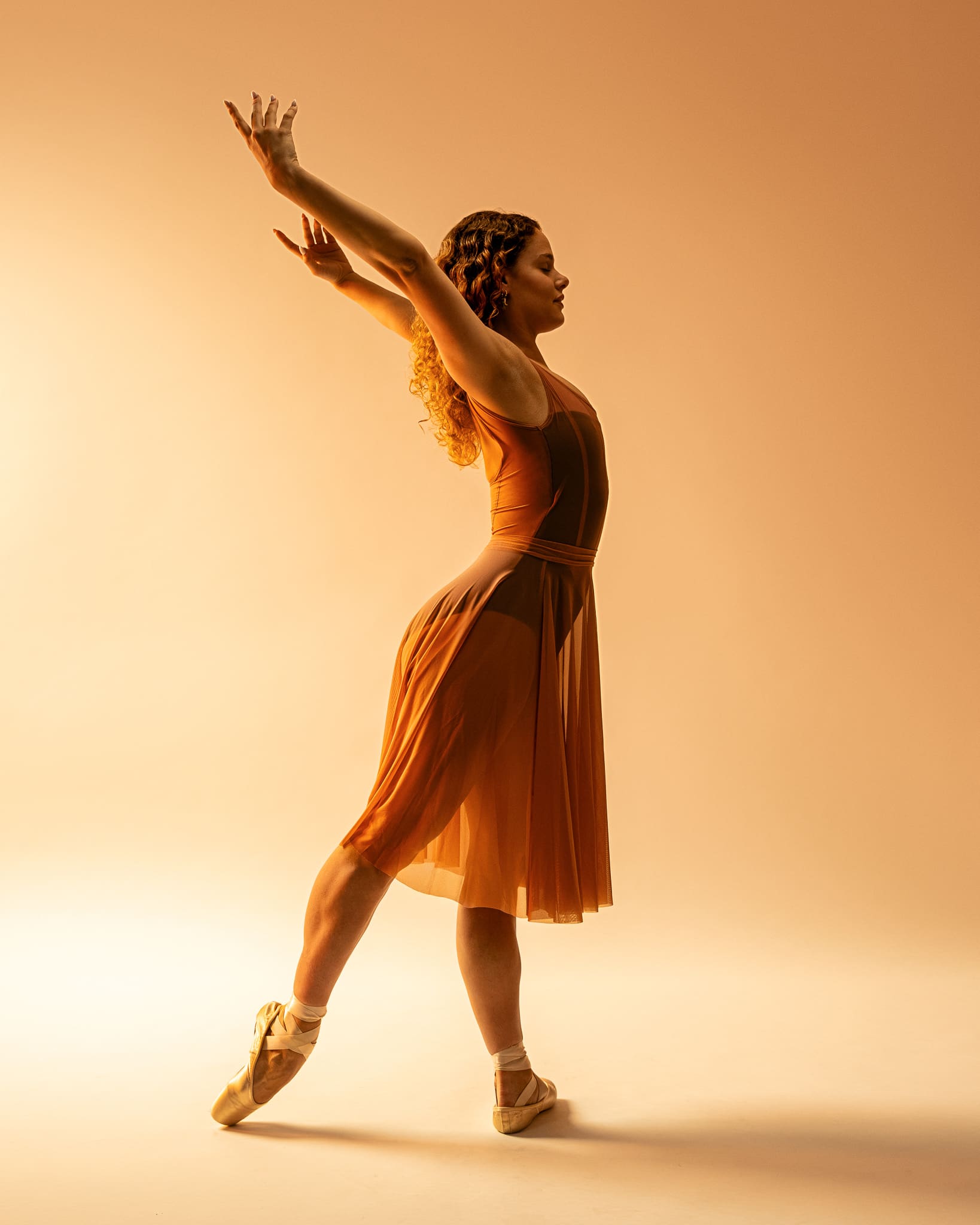Ballet dancer on pointe in a flowing orange dress with one arm raised overhead, warm golden lighting, San Antonio dance photography.