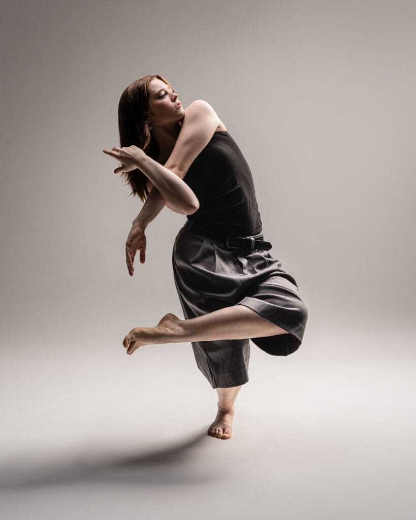 Dancer balanced on one leg with the other bent behind her and arms crossed in front on a neutral backdrop. Photographed by a San Antonio contemporary dance photographer.