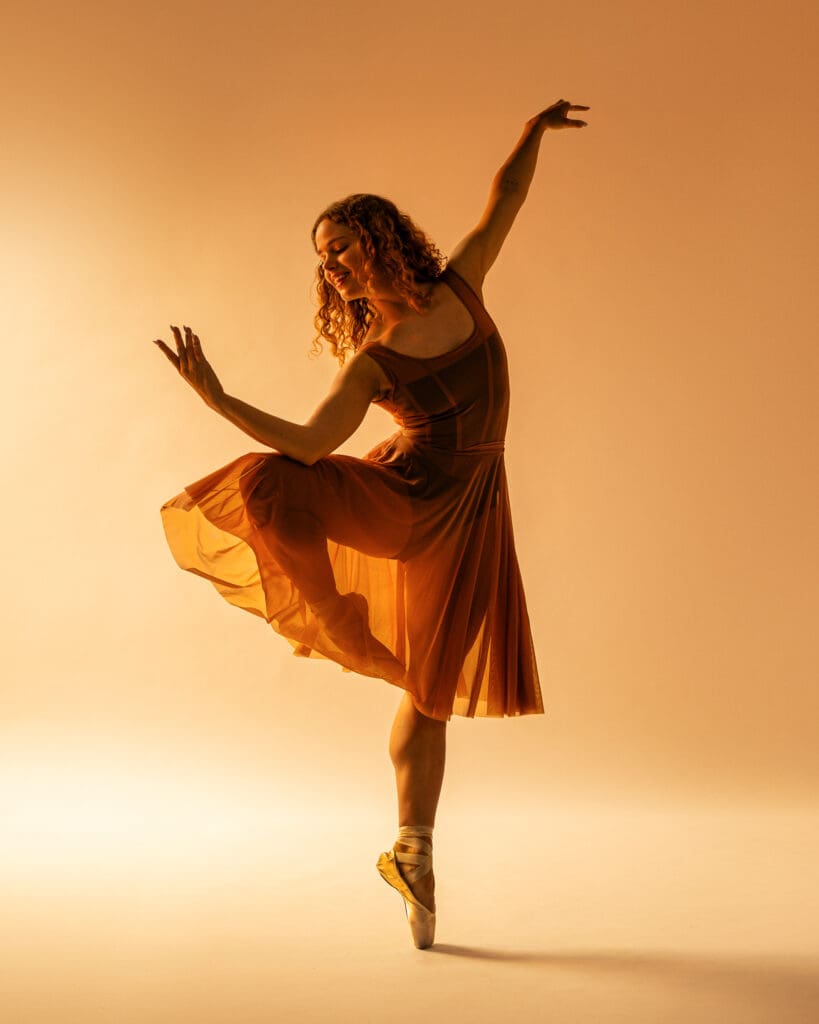 Ballet dancer smiling on pointe in a passé position with arms curved gracefully, warm amber lighting, San Antonio dance photography.