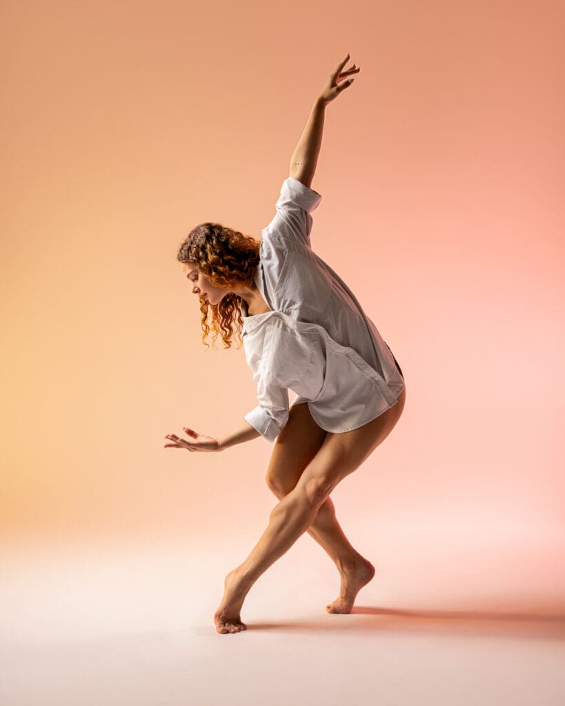 Dancer barefoot in an oversized white shirt, legs crossed and one arm raised overhead, warm peach lighting, San Antonio dance photography.
